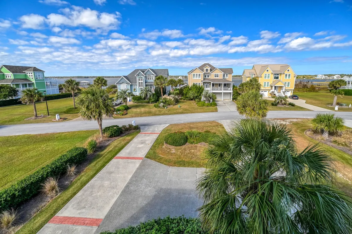 This exterior/view image captures a cheerful neighborhood scene with colorful houses, palm trees, a manicured lawn, and a spacious driveway. The open, sunlit setting suggests vibrant community life, outdoor gatherings, and the broader island environment guests can discover.