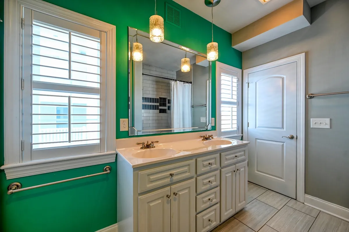 A bright and airy ensuite bathroom with a double vanity sink, large mirror, green accent wall, and modern hanging light fixtures. The design emphasizes both style and function, creating a rejuvenating space for morning and evening routines.