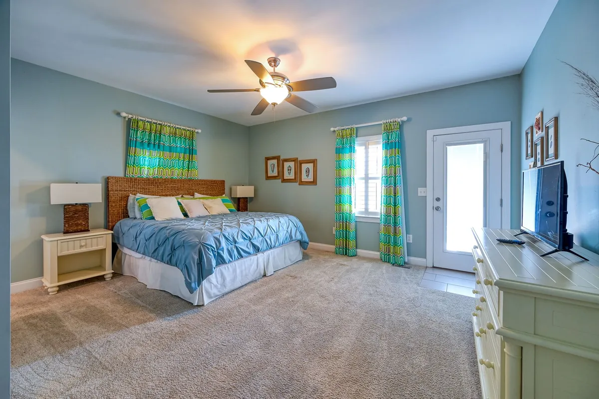 A cheerful bedroom with a bed, curtains, nightstand, television, and door. The space is filled with light and designed for comfort, offering a cozy setting for rest, reading, or enjoying some private downtime.