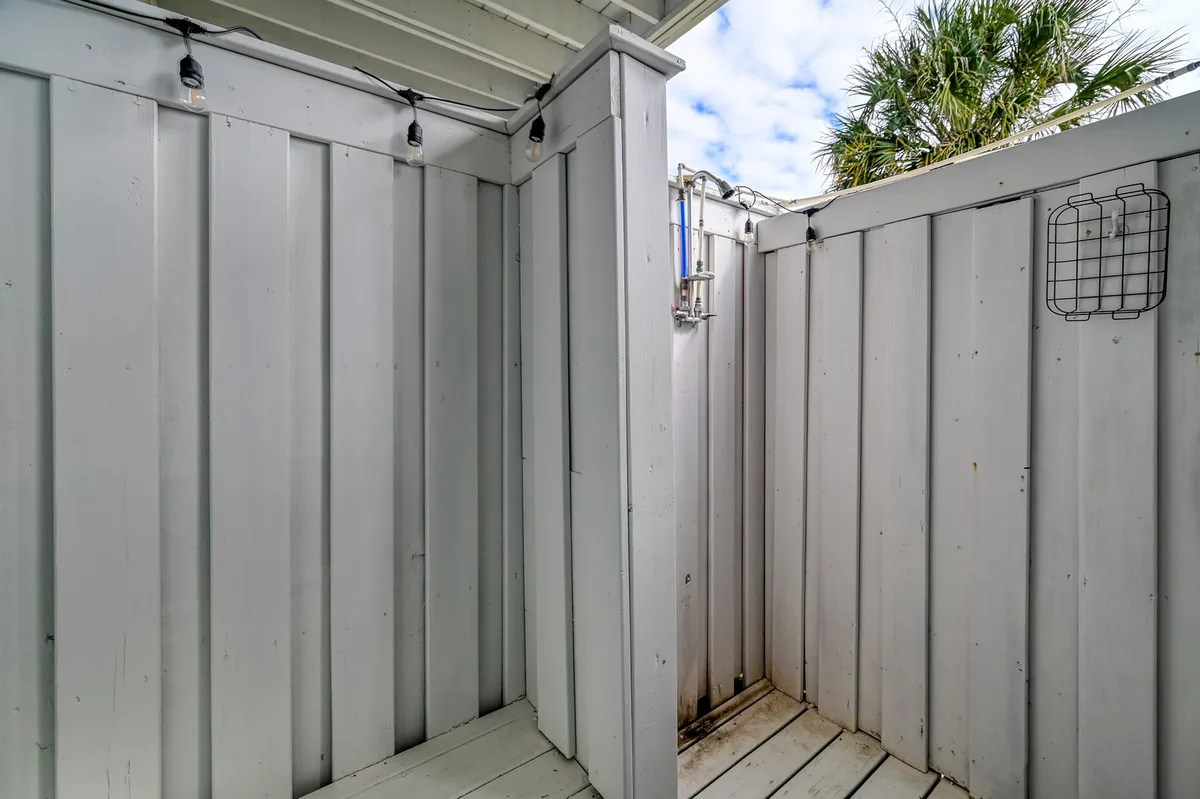 A bright and airy outdoor shower area with wooden slatted flooring, white wooden walls, a modern shower fixture, and decorative string lights. The space feels private yet connected to nature—ideal for a refreshing rinse after a day at the beach or pool.