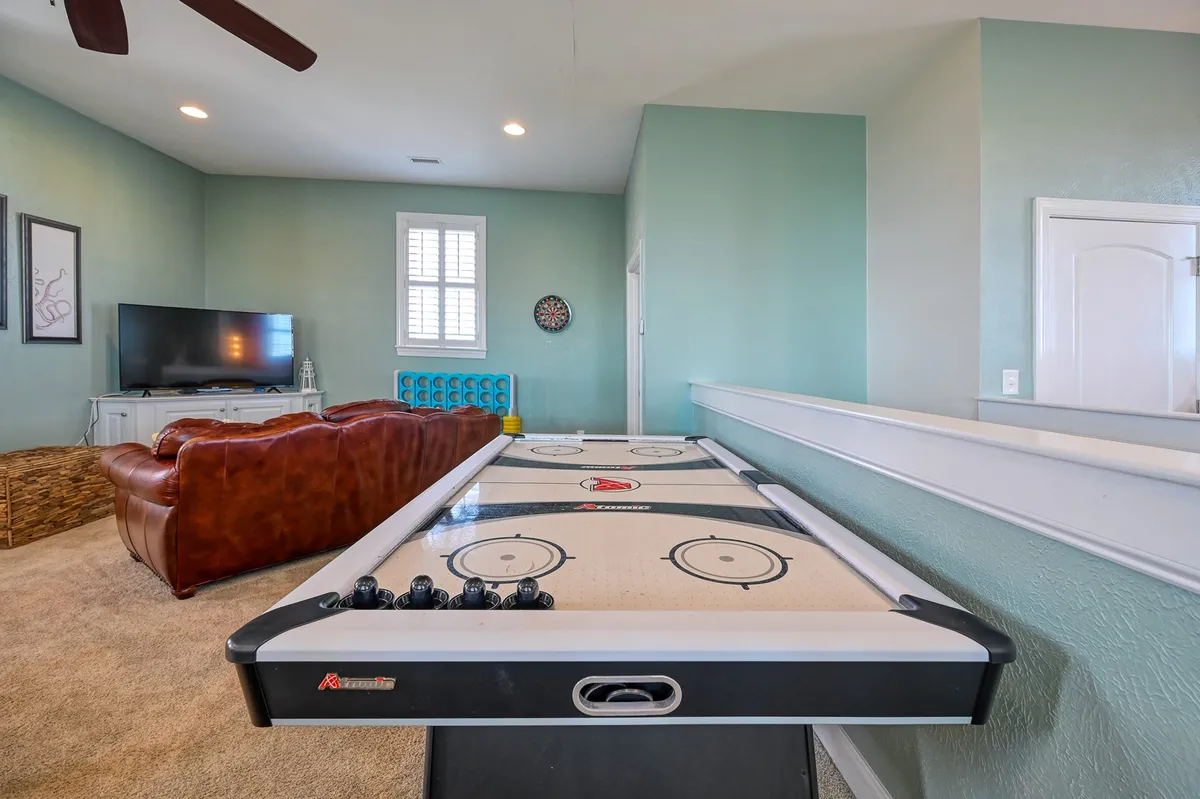 A game room setting with an air hockey table, a comfy brown leather sofa, television, dartboard, and colorful blue seating. The space feels energetic and tailored for teens seeking social interaction and entertainment.
