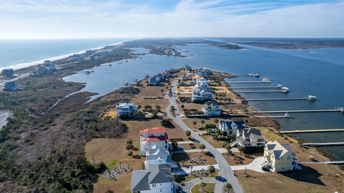 A panoramic view featuring coastal homes, a marina with boats, the sparkling ocean, and a canal winding through the landscape. The scene is bathed in warm natural light, evoking the tranquility and excitement of coastal living and water-based recreation.