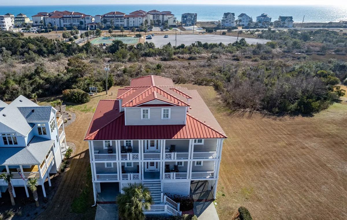 A spacious house with a distinctive red roof sits amid lush greenery, with a glimpse of the ocean in the background. The inviting porch and surrounding landscape evoke a sense of privacy and exclusivity, ideal for relaxation and family gatherings.