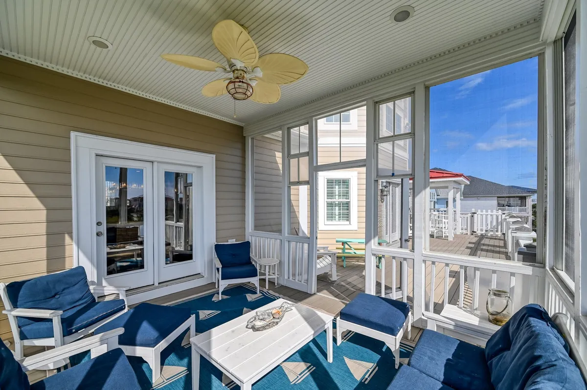A bright, breezy sitting area with blue upholstered chairs, a white coffee table, palm leaf ceiling fan, and large glass doors opening onto a porch. The space invites relaxation and offers access to outdoor breezes and sunset views, connecting indoor living with the porch.
