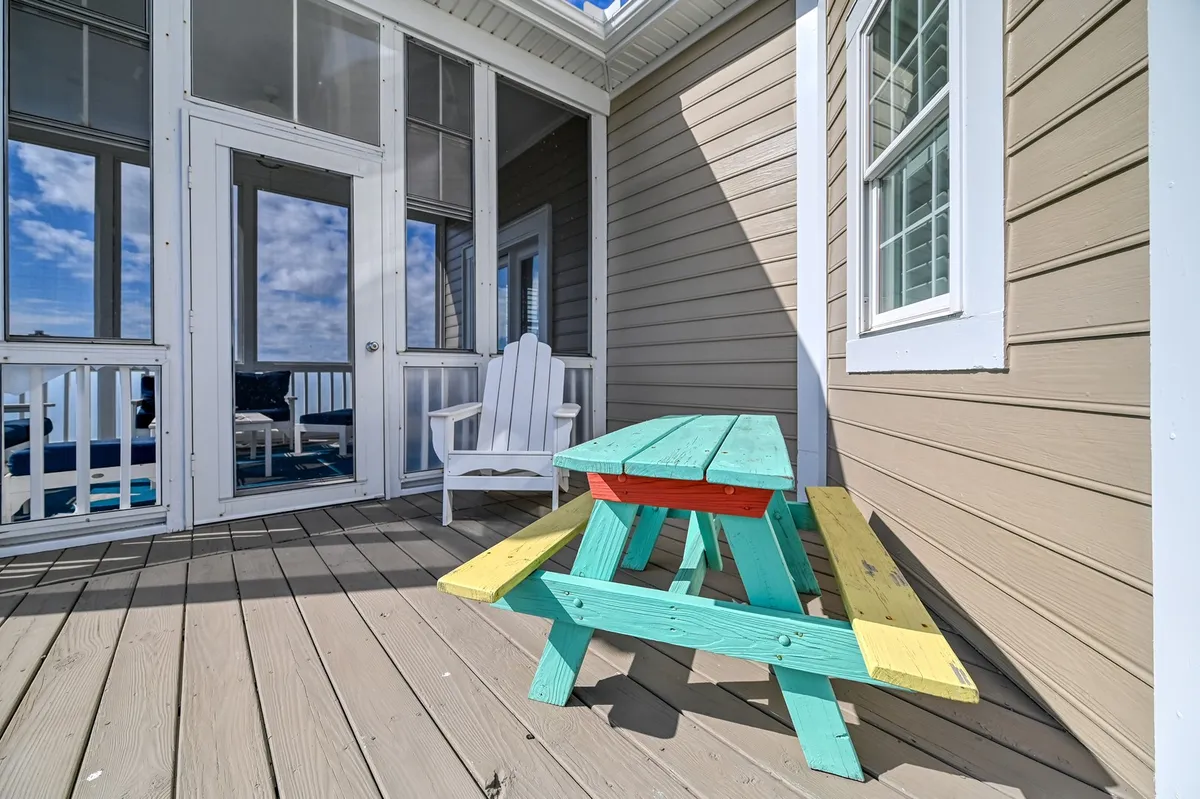 This exterior image features a cheerful setting with a colorful picnic table, a white adirondack chair, and the entrance to a screened porch. The scene radiates a relaxed, social atmosphere with space for outdoor meals, games, and casual gatherings, all set against the inviting backdrop of the home.