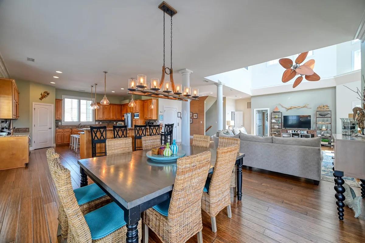 A wide view of a light-filled open-plan interior featuring a large dining table, adjacent open kitchen, cozy living area with a couch, and an eye-catching decorative ceiling fan. The space is styled for gatherings, with plenty of seating and seamless flow between eating, cooking, and relaxing zones.